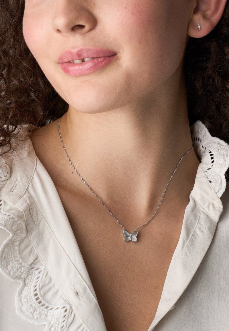 Jonge vrouw met een zilveren vlinderhanger, kleine zilveren oorbel en een witte blouse met een kanten kraag.