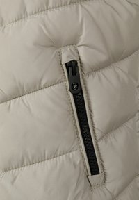 Beige puffer väst med ett diagonal quiltat mönster, en siddragkedja med svarta detaljer och en mjuk, slät tygkänsla.