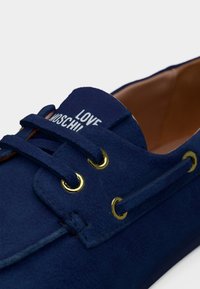 Κοντινό πλάνο ενός μοκασίνι από μπλε σουέντ με χρυσά κρικάκια και τη φράση "LOVE MOSCHINO" στη γλώσσα, που δείχνει το εσωτερικό από δέρμα.