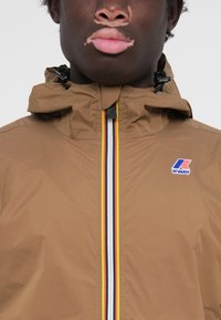 Brown waterproof jacket with a hood, featuring a front zipper with yellow, white, and blue stripes; K-WAY logo on the left side.