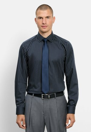 Mann trägt dunkelblaues Nadelstreifenkleidhemd, einfarbige marineblaue Krawatte, graue Hose und schwarzen Gürtel, steht vor schlichtem hellem Hintergrund.