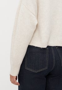 Lys beige genser med ribbet kant og mansjetter, med en løs passform. Brukt sammen med mørkeblå denim jeans med synlige sømdetaljer.