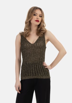Donna con capelli biondi ricci che indossa un top in maglia metallica senza maniche e pantaloni neri, posa con una mano sul fianco su sfondo bianco.