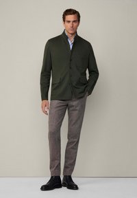 Giacca verde oliva con colletto alto, due tasche frontali e chiusura a bottoni, indossata sopra pantaloni in tweed marrone e stivali in pelle nera.