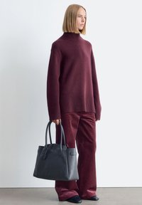 Burgunderfarbener Strickpullover mit geripptem Saum, passende Cordhosen und eine strukturierte schwarze Ledertasche, die über zwei Griffe verfügt und keine sichtbaren Beschläge hat.