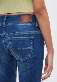 Modre jeans hlače iz jeansa imajo ukrivljen žep na zadnji strani s rumenim šivom. Na pasu je vidna rjava usnjena značkasta nalepka.