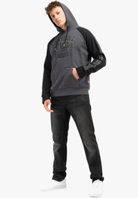 Felpa grigia con accenti neri e logo sul davanti, abbinata a jeans scuri e sneakers bianche, con tasca a marsupio e polsini a costine.
