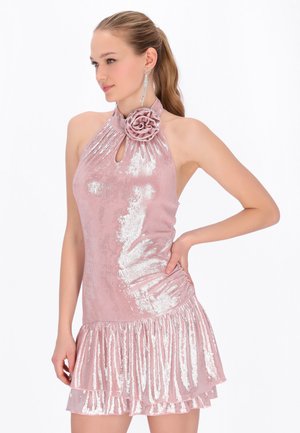 Vrouw draagt een glanzende roze halterjurk met een bloemendetail en een gerimpelde zoom, poseert met één hand op haar heup tegen een witte achtergrond.