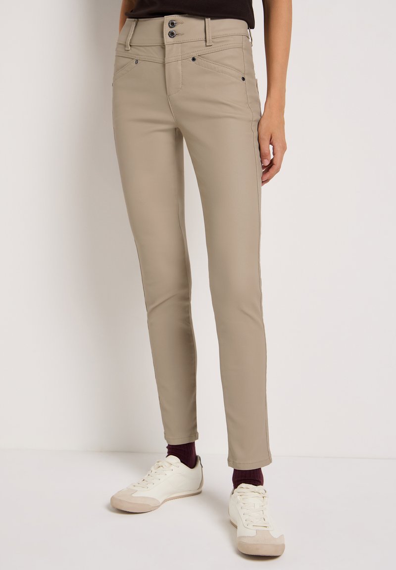 Pantalones ajustados beige con una textura suave, dos bolsillos frontales y cierre de dos botones. Combinados con zapatillas blancas y calcetines burdeos.