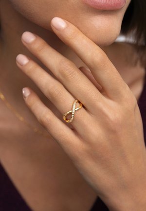 Kultainen äärettömyys-sormus, jossa kaareva sanka ja upotetut kirkkaat jalokivet, esittäen sileää tekstuuria ja kiillotettua pintaa.