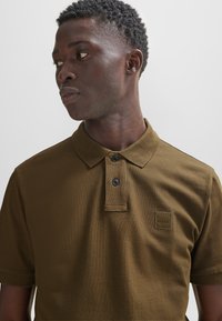 Braunes Poloshirt mit zwei schwarzen Knöpfen und einem Patch-Label auf der linken Brust. Texturiertem Stoff und klassischem Kragen-Design.