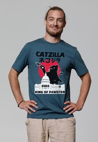 T-shirt en coton teal avec un design graphique représentant un grand chat sur le bâtiment du Capitole, avec le texte "CATZILLA" et "KING OF PAWSTER".