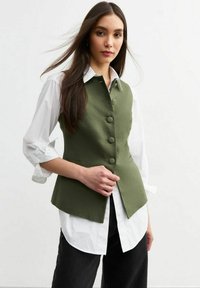 Chaleco sin mangas en verde olivo con seis botones redondos, llevado sobre una camisa blanca. Tejido ligero con una textura suave y una forma estructurada.