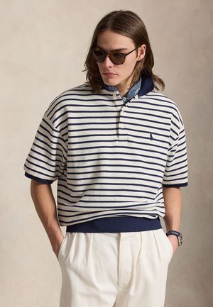 Ung mand iført skildpaddemønstret solbriller, marineblå og hvide stribede poloshirt, blå mønstret tørklæde, hvide bukser, med hænderne i lommerne.