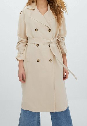 Femme portant un trench-coat beige à double boutonnage avec une ceinture nouée à la taille et un jean bleu à jambes larges sur un fond uni.