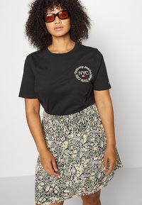 T-shirt de algodão preto com o logo "Tommy Jeans NYC", combinado com uma saia floral com padrões verdes, rosas e amarelos sobre um fundo preto.