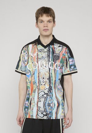 Junger Mann, der ein kurzärmeliges, mehrfarbiges Hemd mit schwarzem Kragen und "Colucci"-Logo trägt, vor einem schlichten hellen Hintergrund steht.