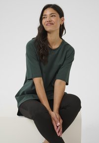 Donker groene oversized T-shirt met korte mouwen en een ronde halslijn, gecombineerd met strakke zwarte broeken. Gladde textuur en casual ontwerp.