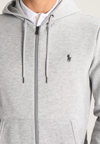 Grå zip-up hoodie i mjukt tyg. Har en framzipper, dragkedje snoddar, en logotyp på bröstet och två framfickor.