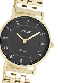 Montre dorée avec un cadran noir, des index romains et un bracelet en métal lisse. Dispose d'une couronne simple et de l'étiquette "OOZOO Vintage".