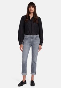 Blusa nera con maniche a sbuffo e dettagli in pizzo, abbinata a jeans grigi chiari con risvolti e mocassini neri. Il modello si trova su uno sfondo neutro.