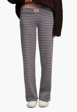 Femme portant un pantalon long à jambes larges avec des rayures horizontales bleues, marron et blanches, des baskets blanches et un pull marron foncé.