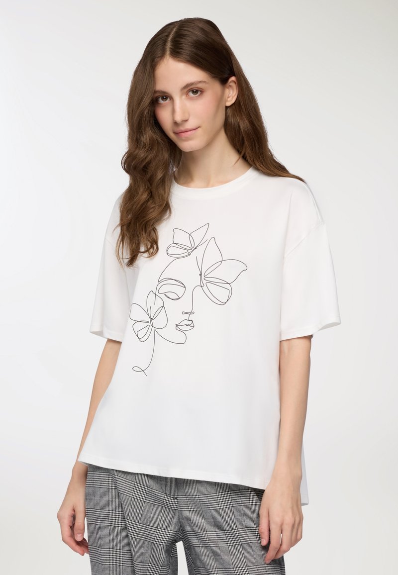 Biała koszulka z krótkim rękawem z czarnym rysunkiem twarzy i motyli. Gładka tekstura, luźny krój, w połączeniu z szarymi spodniami w kratę.