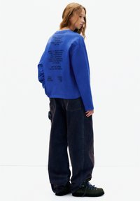 Hanorac albastru cu text negru pe spate, asortat cu pantaloni oversized din denim închis și bocanci verzi de drumeție. Textură netedă a țesăturii.