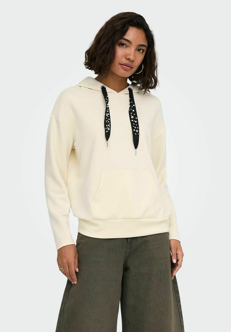 Femme portant un sweat à capuche écru avec des cordons noirs ornés et un pantalon ample vert foncé, debout sur un fond clair uni.