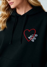 Felpa nera con cappuccio caratterizzata da un design ricamato a forma di cuore rosso e spille di sicurezza bianche. Tessuto morbido, cappuccio con cordino e tasca frontale.