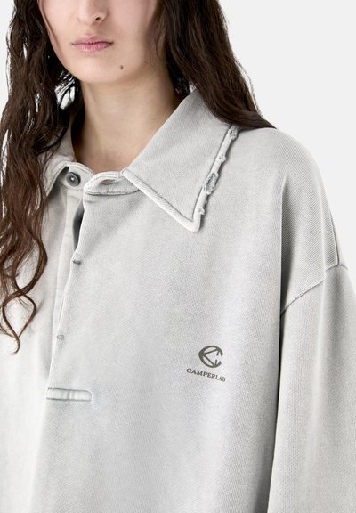 Femme portant un polo gris clair avec des bords de col effilochés et un petit logo « Camperlab » sur le côté gauche de la poitrine.
