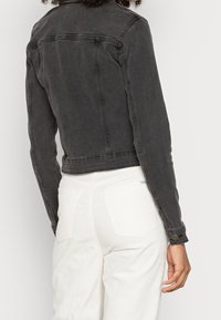 Chaqueta de mezclilla negra con una silueta entallada, que presenta mangas largas, puños de botón y costuras visibles. Llevada sobre pantalones de pana blancos.