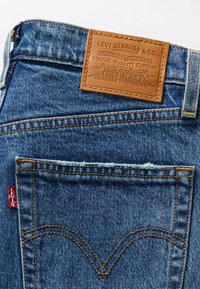 Poche arrière d'un jean en denim bleu avec un patch en cuir marron Levi Strauss & Co. et une petite étiquette en tissu rouge Levi's sur la couture latérale.