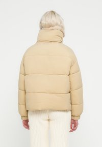 Μπεζ μπουφάν puffer με ψηλό γιακά, καπιτονέ σχέδιο και ελαστικές μανσέτες, συνδυασμένο με λευκό πλεκτό παντελόνι με υφή.