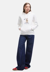 Jongeman in een witte hoodie met een konijn en de tekst "Happy", wijde blauwe jeans en rode sneakers, staand met handen in de zakken.