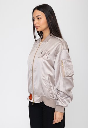 Kobieta o długich ciemnych włosach ubrana w jasnobeżową satynową kurtkę bomber z wyszywanym literą „B” i kieszenią na zamek na rękawie.