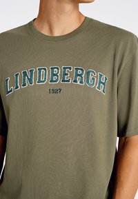 Olivgrön bomullströja med rund halsringning och korta ärmar. Har fet, broderad text "LINDBERGH" och "1927" under.