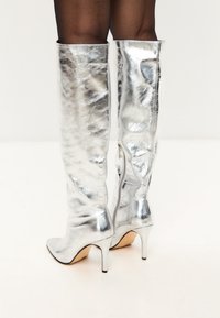 Ghete metalice argintii până la genunchi, cu vârf ascuțit și toc stiletto, având o textură elegantă și netedă, cu închidere prin fermoar la spate.