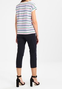 Kvinna i en kortärmad, flerfärgad randig skjorta, avklippta svarta byxor och svarta högklackade ankelsandaler, stående med ryggen vänd.