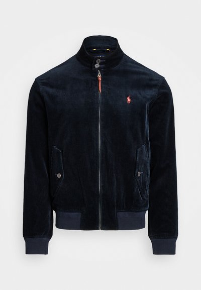 Giacca nera in corduroy con zip, colletto alto, tasche con bottone, polsini e vita a coste, e logo ricamato rosso sul petto.