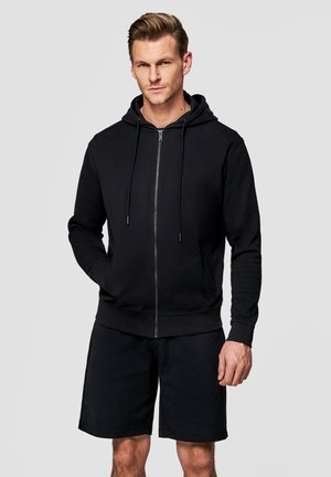 Mężczyzna o blond włosach, ubrany w czarną bluzę z zamkiem i czarne szorty, stoi z jedną ręką w kieszeni na jednolitym tle.