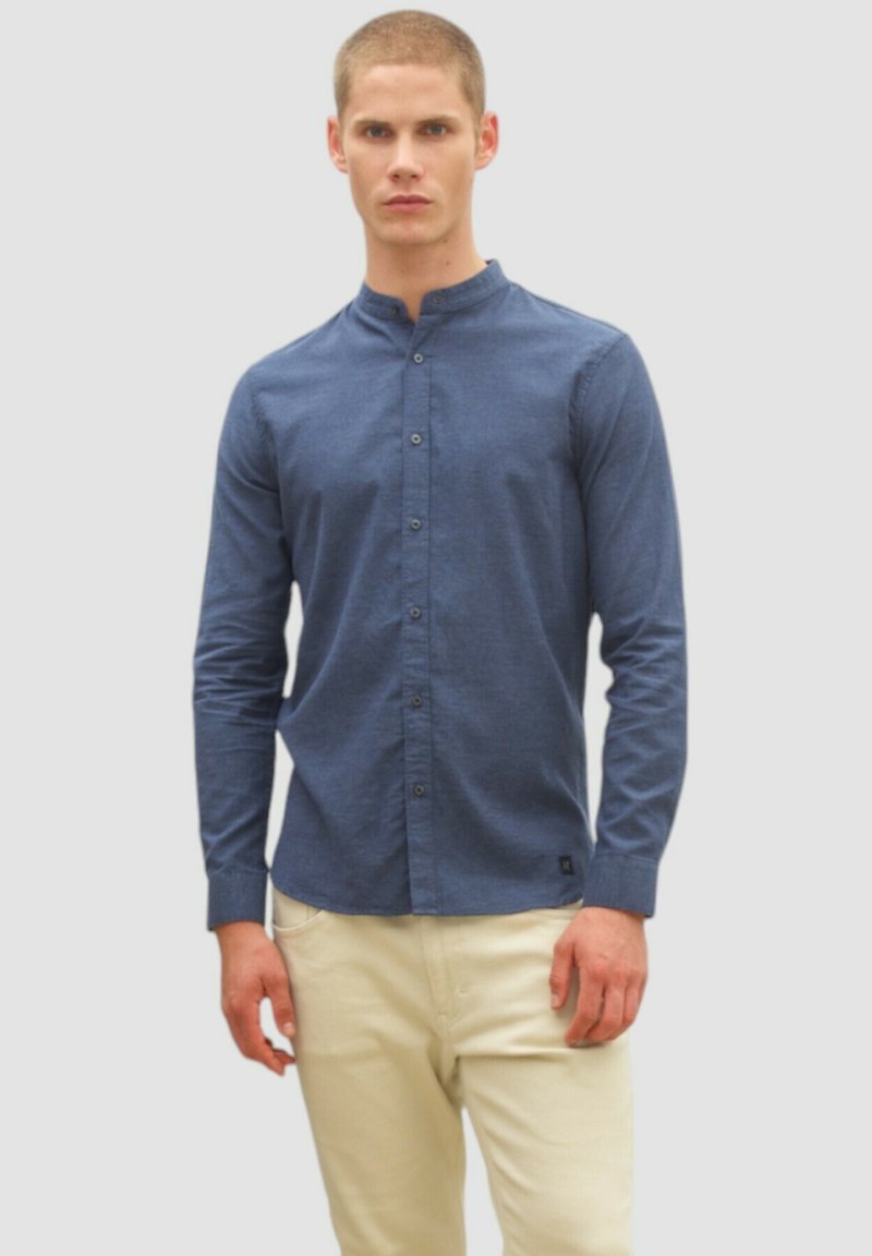 Langarm-Hemd aus navyblauem Stoff mit Stehkragen, vorderem Knopfverschluss und schmaler Passform. Kombiniert mit hellbeigen Hosen.