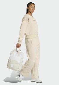 Tuta beige con dettagli a coste, vestibilità oversize e polsini elastici. Tiene uno zaino bianco e beige chiaro. Le sneakers sono bianche con dettagli neri.