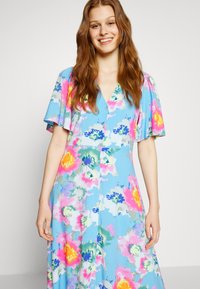 Vestido floral em tecido azul com padrões coloridos em rosa, amarelo e verde. Decote em V, mangas curtas com babados e detalhe de botões à frente.