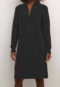 Femme portant une robe en maille noire à manches longues avec un col zippé et des fentes latérales, debout devant un fond clair uni.