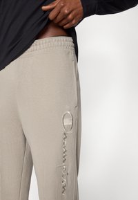 Φωτεινό μπεζ παντελόνι φόρμας με ελαστική μέση, με κεντημένο το υφασμάτινο λογότυπο "Champion" σε πιο ανοιχτή α πόχρωση στο πλάι.