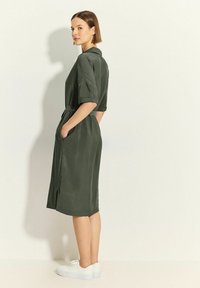 Abito camicia verde oliva con maniche corte, vita con cintura, tasche laterali e dettaglio con spacco; abbinato a sneaker bianche. Tessuto dalla texture liscia.