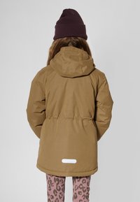 Veste matelassée marron avec capuche, taille cintrée et écusson logo blanc. Assortie à des leggings imprimé léopard rose. Personne vue de dos.