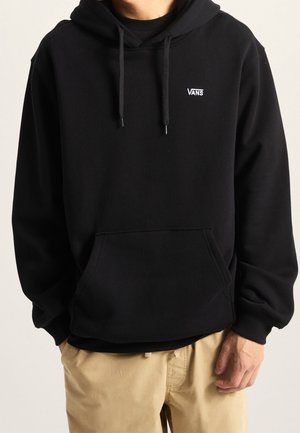Personne portant un sweat à capuche noir Vans avec cordons et une poche avant, associé à un pantalon beige, debout devant un fond neutre.