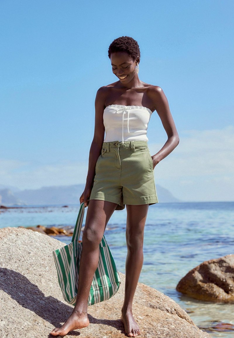 Femme debout pieds nus sur des rochers au bord de la mer, portant un haut tube blanc, un short vert et tenant un sac fourre-tout rayé vert.